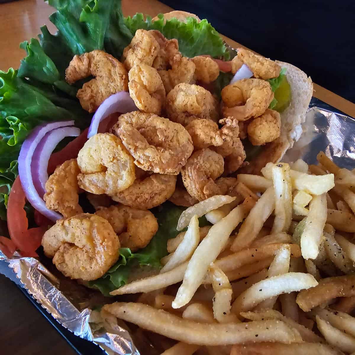 Fried shrimp po'boyw with lettuce, tomatoes, and red onions next to fries in a basket. 