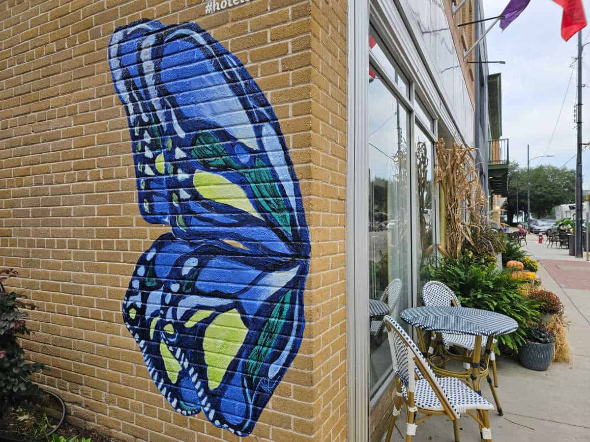 Colorful blue butterfly wing mural on the side of a brick building next to table and chairs at the hotel entrance