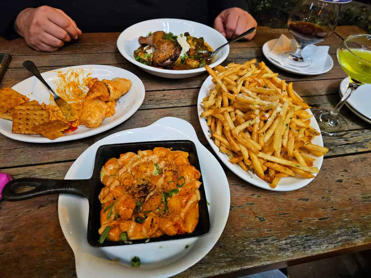 White plates with food on a picnic table outside. Hands and drinks on the edges of the table