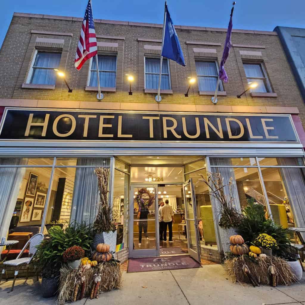 Hotel Trundle sign over glass windows and entrance door of a brick building with fall decorations on either side of the door, three flags hanging above the hotel trundle sign