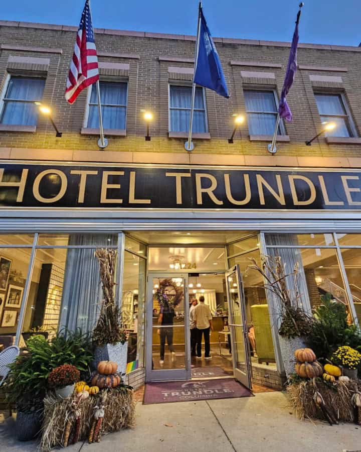 Hotel Trundle sign over glass windows and entrance door of a brick building with fall decorations on either side of the door, three flags hanging above the hotel trundle sign