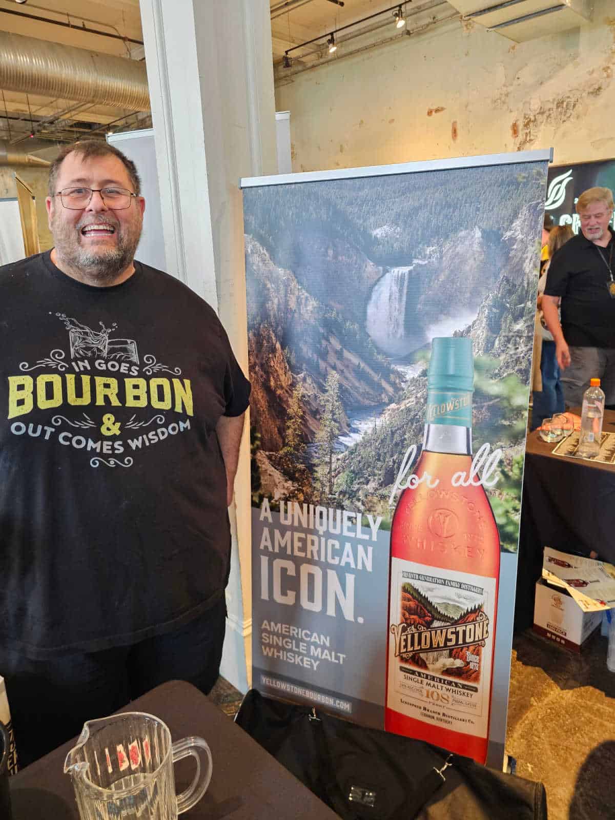 John next to a Yellowstone Bourbon sign with Yellowstone falls in the background. 