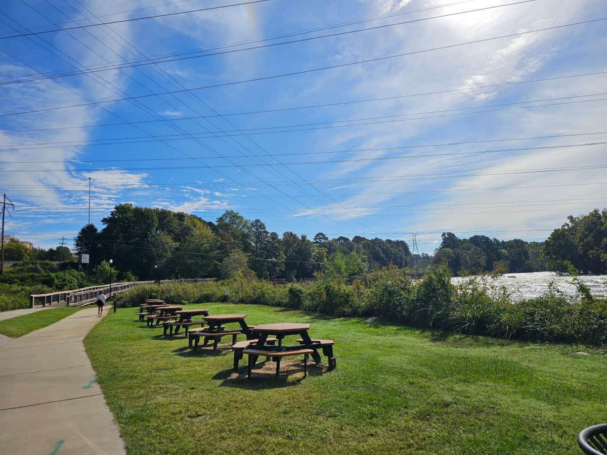 picnic tables lined up in a grassy area with a walkway next to them and the Saluda river on the other side. 